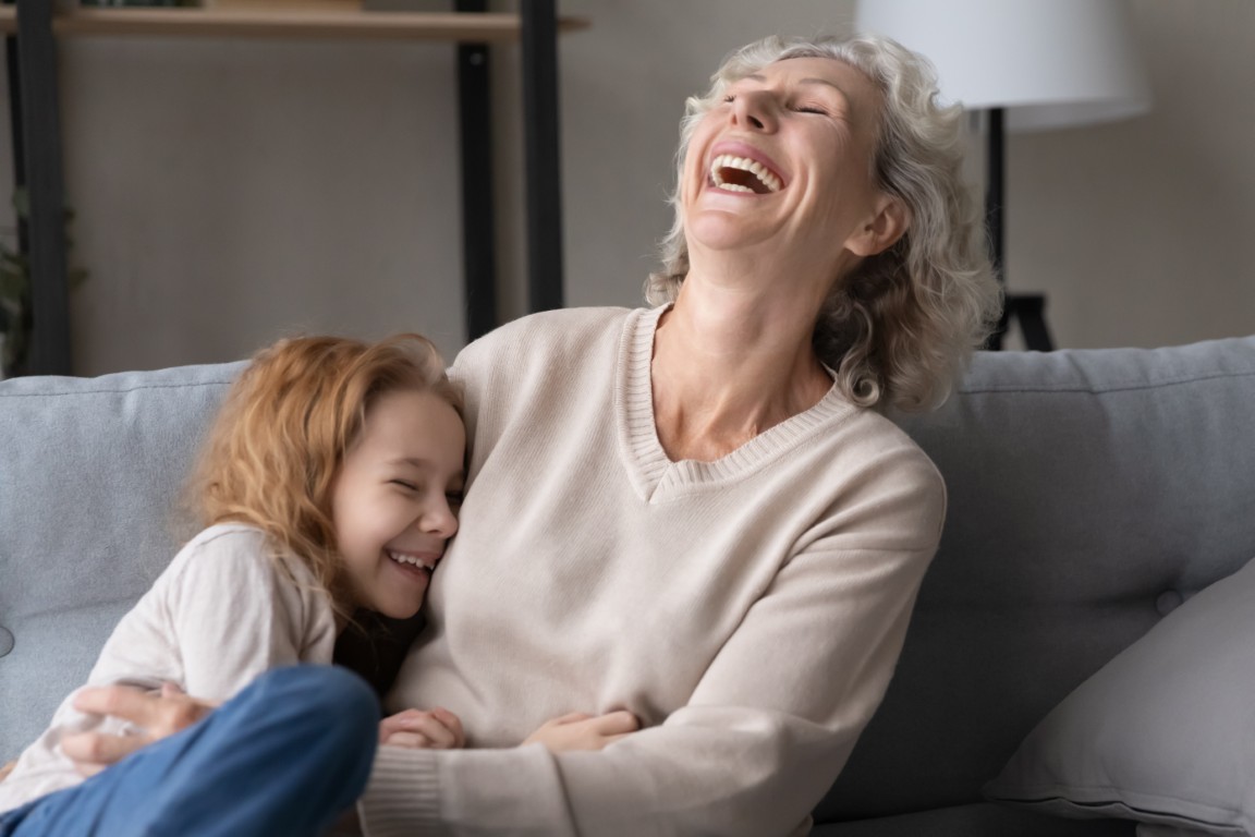 A grandmother laughs while cuddling her granddaughter, they're both sat on the sofa